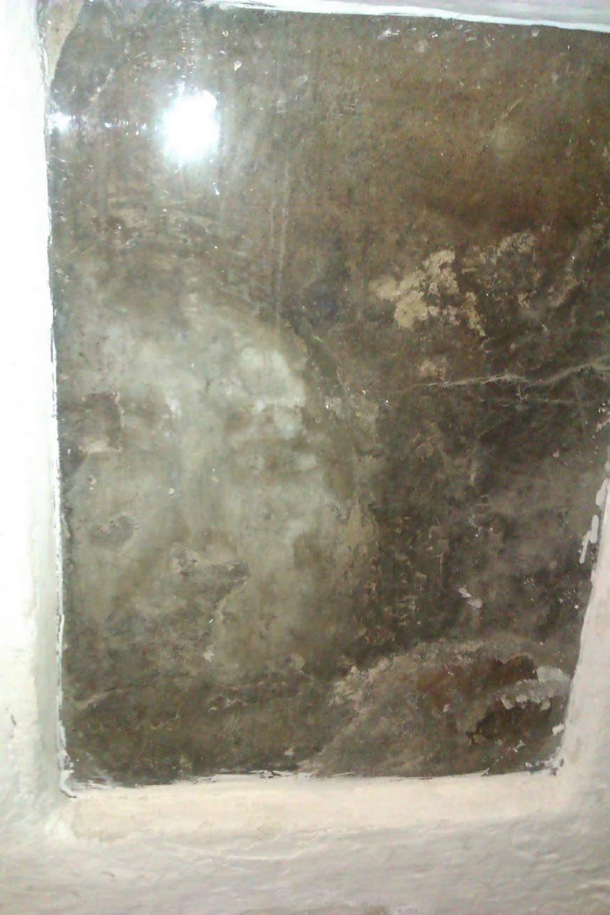 Mancha oscura con forma de rostro en una losa de cemento, tras un cristal con el reflejo de un flash