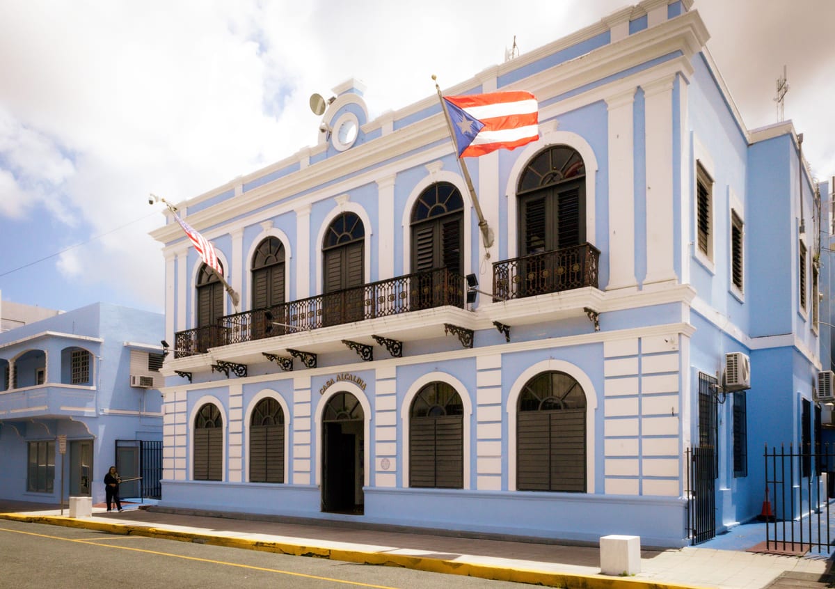 Fachada azul y blanca de la casa alcaldía de Canóvanas, con la bandera de Puerto Rico en el balcón