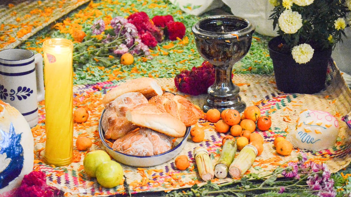 Ofrenda sobre un petate con pan, tejocotes, cañas de azúcar, una vela, un sahumerio y una calavera de azúcar