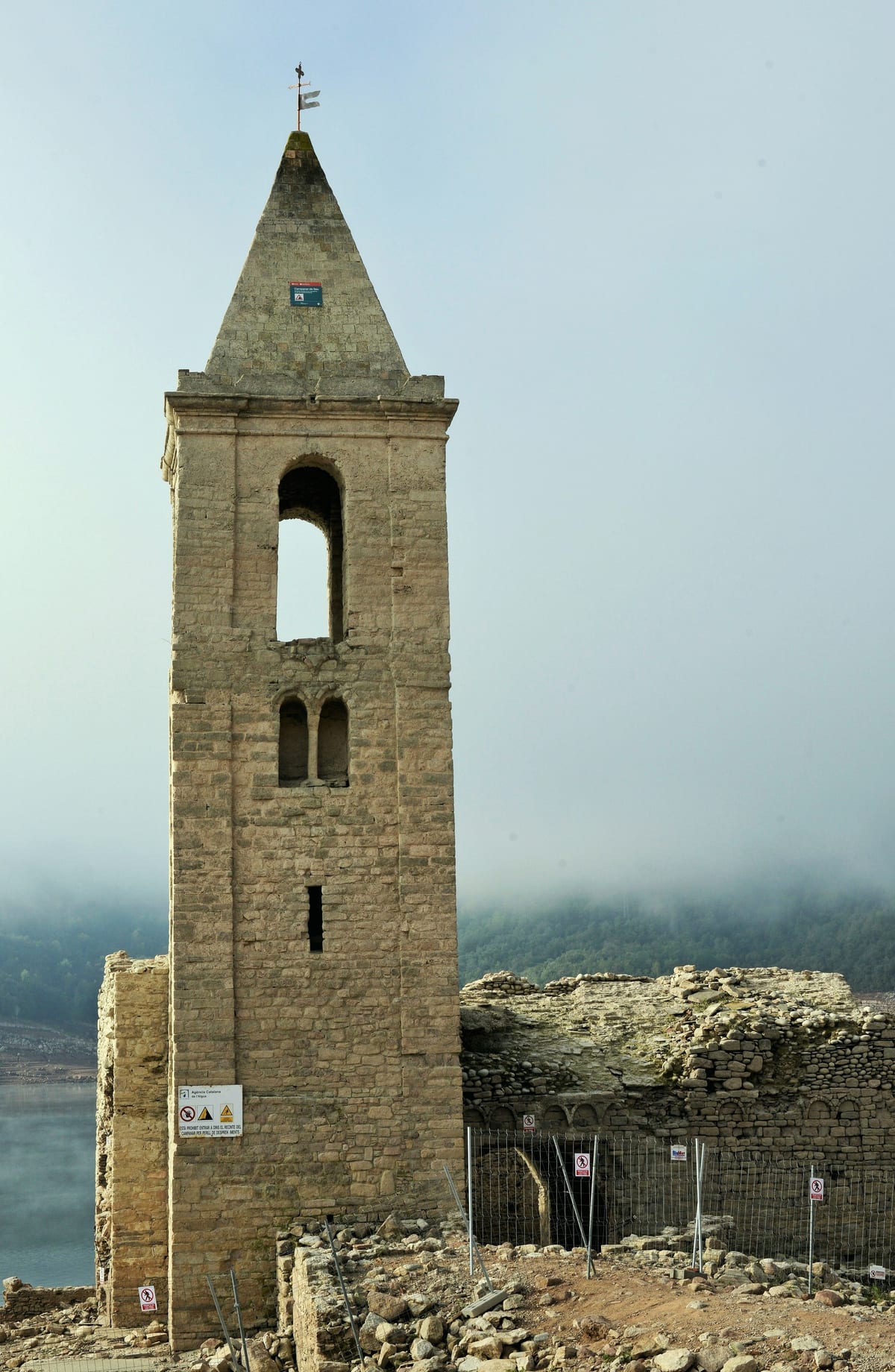 El campanario de piedra de Sant Romà de Sau, fuera del agua, con un cartel de prohibido entrar y vallas metálicas