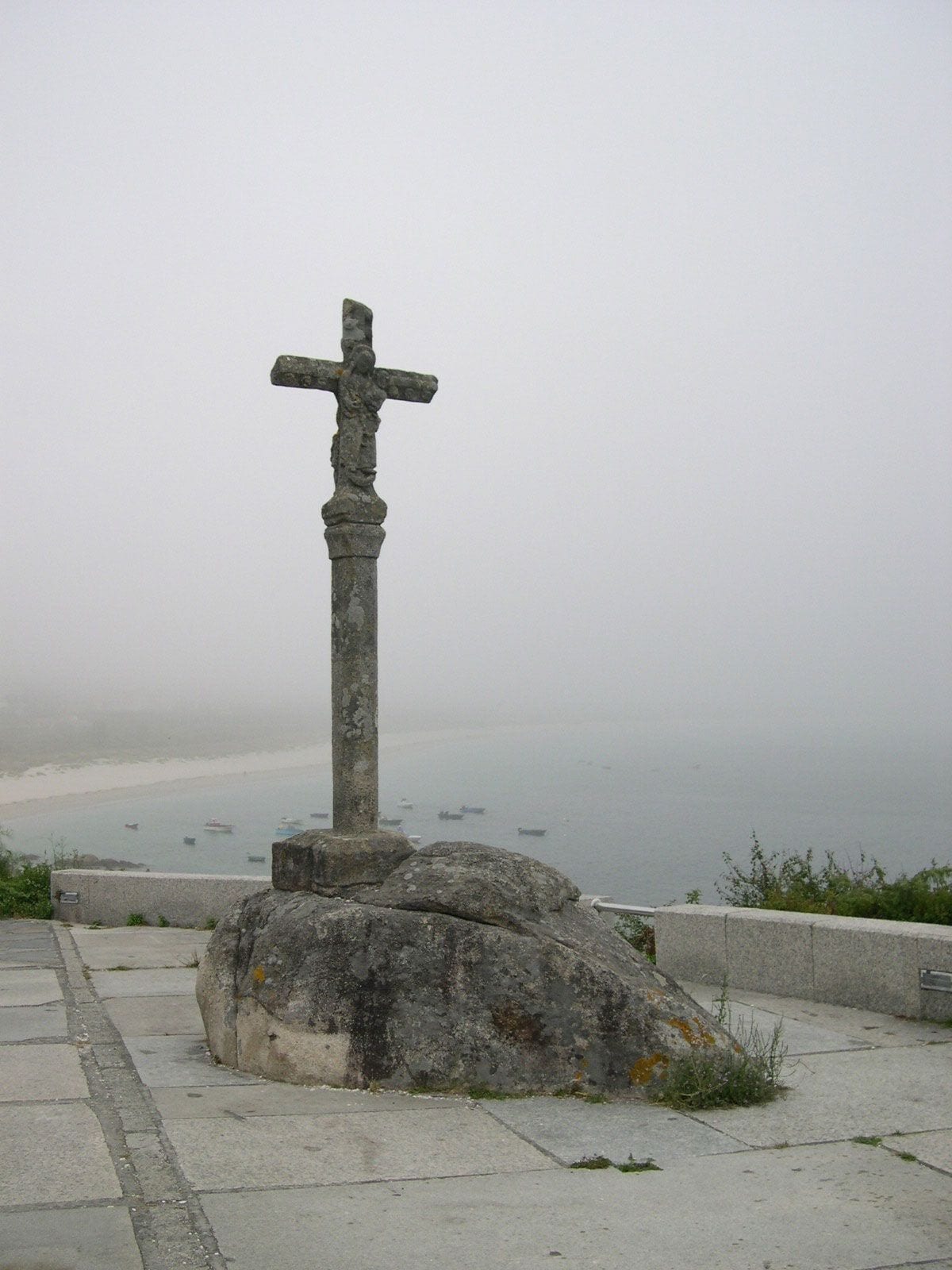 Cruz de piedra sobre un fuste alto y una roca, con la niebla tapando el mar y una playa con barcas